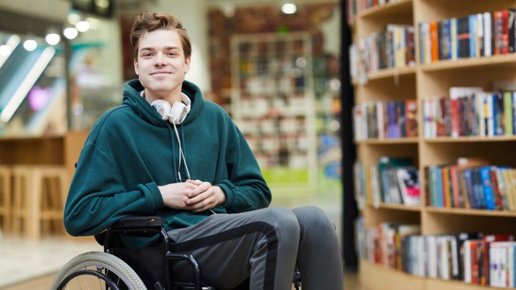 Young man with brown hair wearing headphones around his neck, dark green hoodie and grey sweatpants with a stripe. He sits in a wheelchair looking at the camera with his hands on his lap.