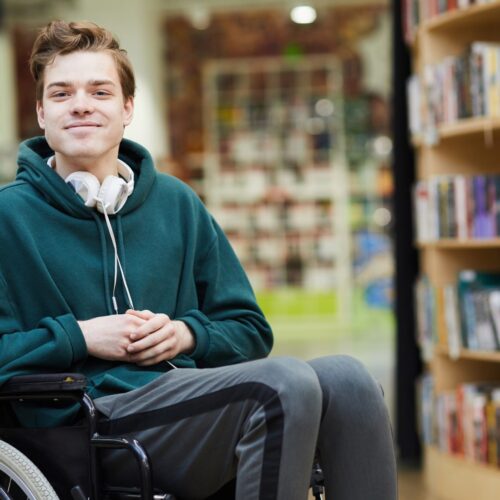 Young man with brown hair wearing headphones around his neck, dark green hoodie and grey sweatpants with a stripe. He sits in a wheelchair looking at the camera with his hands on his lap.